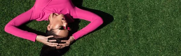 Colpo panoramico di donna attraente in abito e occhiali da sole in posa e sdraiato sull'erba al di fuori — Foto stock