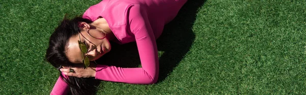 Tiro panorâmico de mulher atraente em vestido e óculos de sol posando e deitado na grama fora — Fotografia de Stock