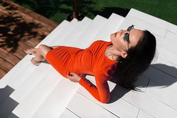 Vue grand angle de la femme attrayante en robe et lunettes de soleil posant et assis sur les escaliers à l'extérieur — Photo de stock