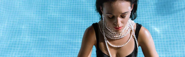 Panoramic shot of attractive woman in black swimsuit and pearl necklace posing in pool — Stock Photo