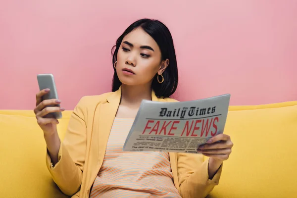 Serious asian woman holding newspaper with fake news and smartphone on yellow sofa isolated on pink — Stock Photo