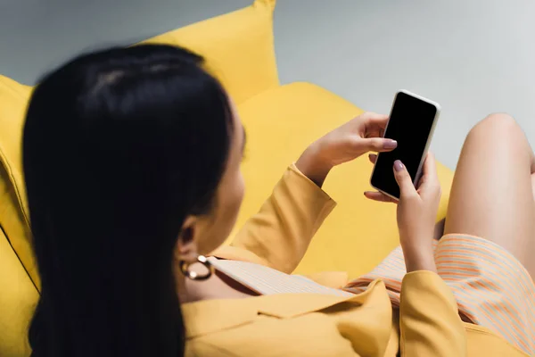 Overhead view of asian woman holding smartphone while sitting on yellow couch isolated on grey — Stock Photo