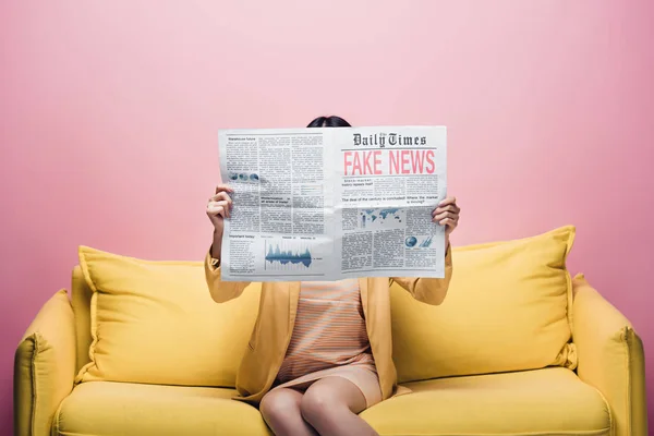 Asian woman holding newspaper with fake news in front of face while sitting on yellow sofa isolated on pink — Stock Photo