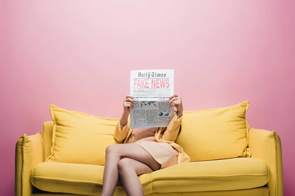 Азиатка держит газету с фейковыми новостями перед лицом, сидя на желтом диване, изолированном на розовом — стоковое фото