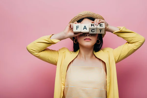Asian woman holding wooden cubes in front of face with fake and fact lettering isolated on pink — Stock Photo