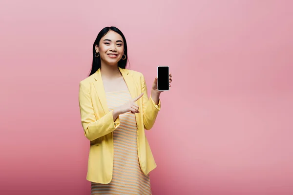 Glücklich asiatische Frau in gelbem Outfit zeigt mit dem Finger auf Smartphone mit leerem Bildschirm isoliert auf rosa — Stockfoto