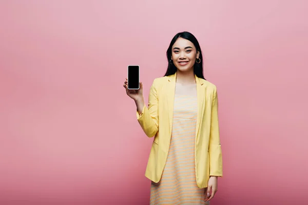 Happy asian woman in yellow outfit holding smartphone with blank screen isolated on pink — Stock Photo