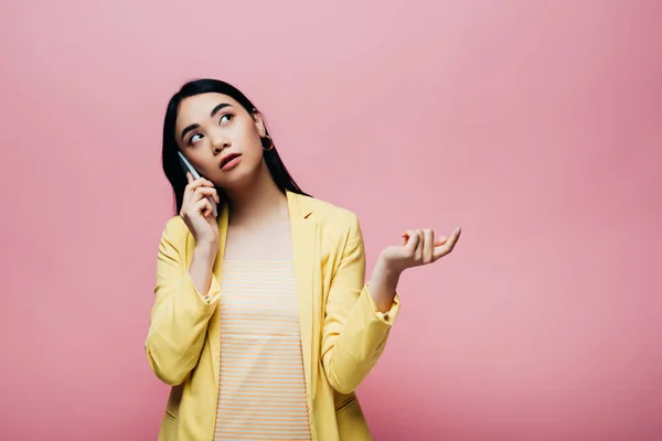 Soñadora asiática mujer en amarillo traje hablando en smartphone aislado en rosa - foto de stock