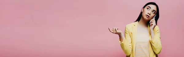 Мечтательная азиатка в желтом наряде разговаривает на смартфоне изолированный на розовый, панорамный снимок — стоковое фото