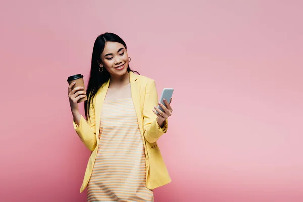 Улыбающаяся азиатка в жёлтом костюме держит бумажную чашку и использует смартфон, изолированный на розовом — стоковое фото