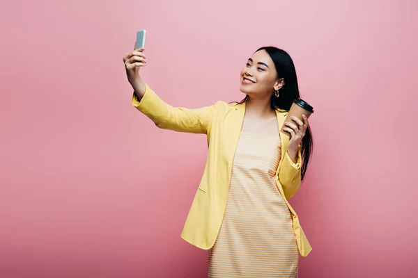 Happy asian woman in yellow outfit holding paper cup and taking selfie isolated on pink — Stock Photo