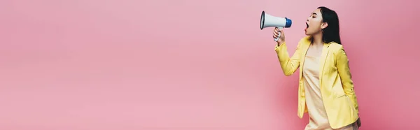 Side view of asian woman in yellow outfit screaming in megaphone isolated on pink, panoramic shot — Stock Photo