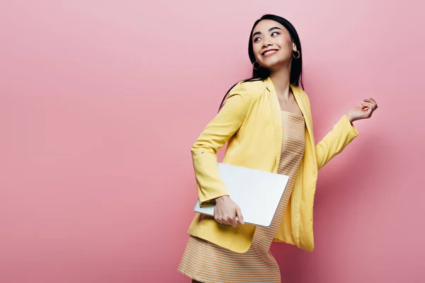Happy asian woman in yellow outfit holding laptop isolated on pink — Stock Photo
