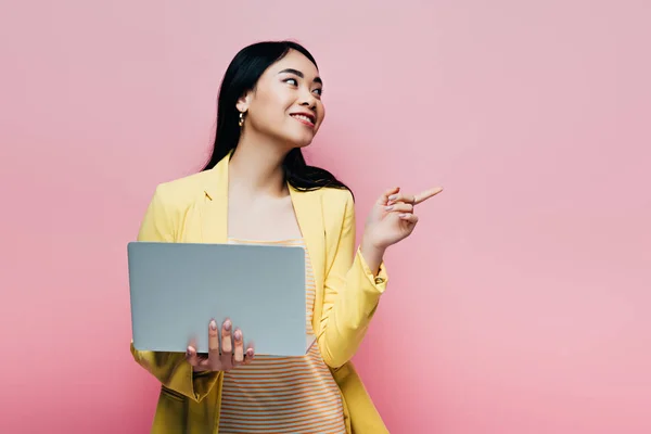Happy asian woman in yellow outfit holding laptop and pointing with finger away isolated on pink — Stock Photo