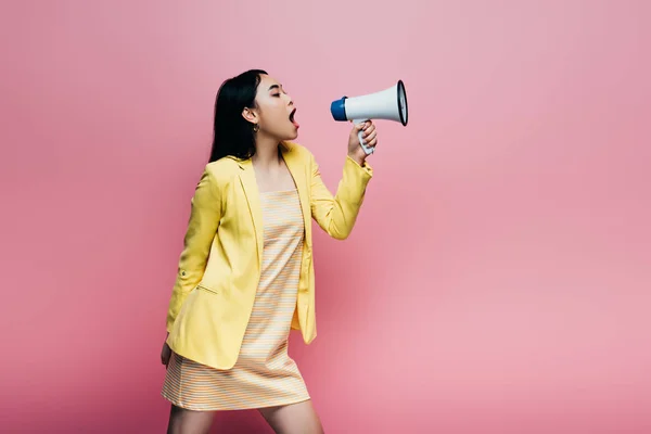 Vista lateral de mujer asiática en traje amarillo gritando en megáfono aislado en rosa - foto de stock