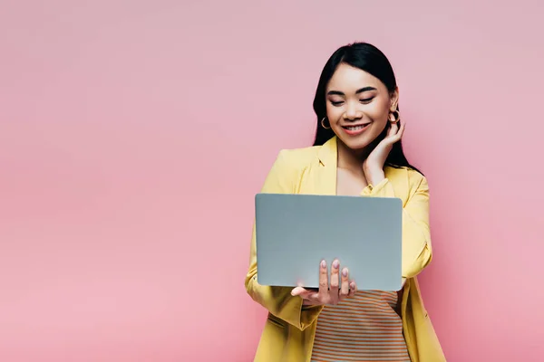 Happy asian woman in yellow outfit holding laptop isolated on pink — Stock Photo