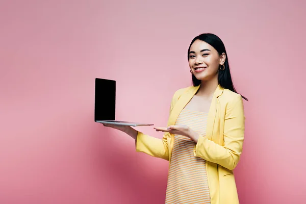 Happy asian woman in yellow outfit holding laptop with blank screen isolated on pink — Stock Photo