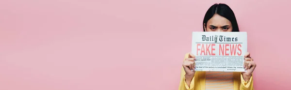 Triste asiático mujer en amarillo traje celebración periódico con falso noticias aislado en rosa, panorámica disparo - foto de stock