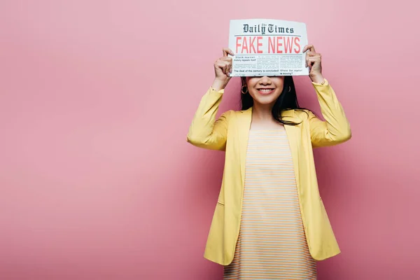 Glücklich asiatische Frau in gelbem Outfit hält Zeitung mit Fake News isoliert auf rosa — Stockfoto