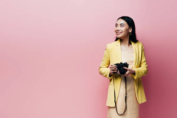 Lächelnde Asiatin im gelben Outfit mit Digitalkamera und isoliertem Blick auf rosa — Stockfoto