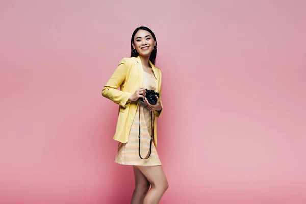 Feliz asiático mujer en amarillo traje celebración digital cámara aislado en rosa - foto de stock