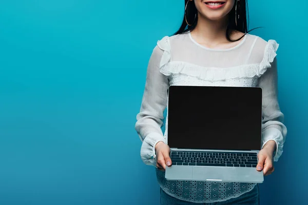 Recortado vista de sonriente mujer asiática en blanco blusa celebración de ordenador portátil con pantalla en blanco sobre fondo azul - foto de stock