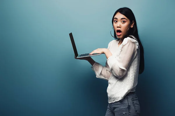 Shocked asian woman in white blouse holding laptop on blue background — Stock Photo
