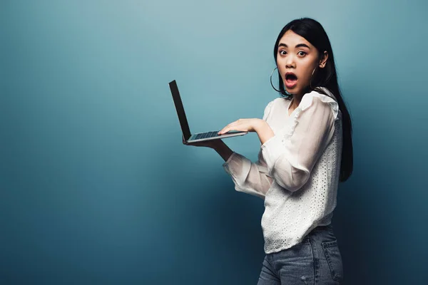 Conmocionada mujer asiática en blusa blanca sosteniendo portátil sobre fondo azul - foto de stock