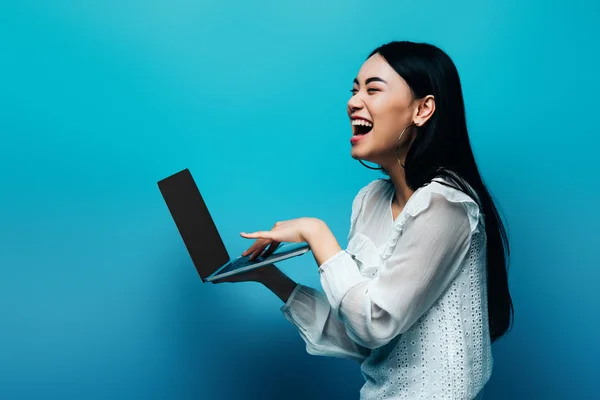 Feliz asiático mujer en blanco blusa holding laptop y riendo en azul fondo - foto de stock