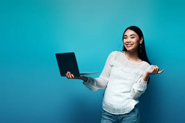 Sonriente asiático mujer en blanco blusa holding laptop en azul fondo - foto de stock