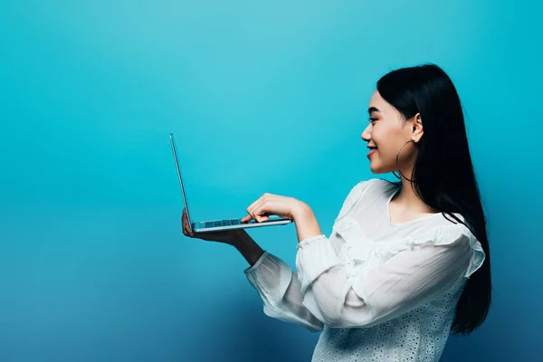 Vista laterale di sorridente donna asiatica in camicetta bianca che tiene il computer portatile su sfondo blu — Foto stock