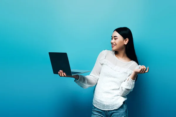 Sonriente asiático mujer en blanco blusa holding laptop en azul fondo - foto de stock