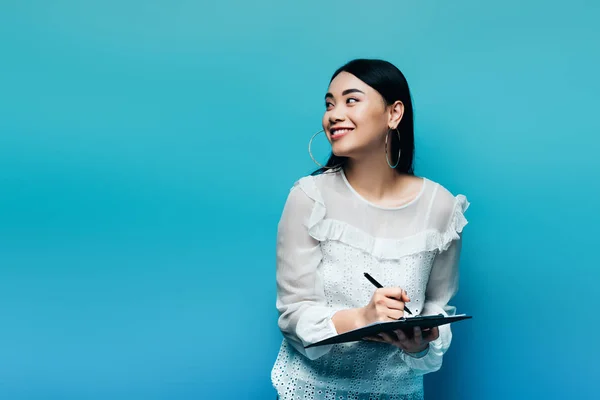 Happy asian journalist in white blouse writing on clipboard on blue background — Stock Photo