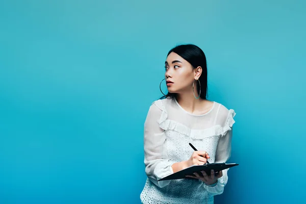 Glücklicher asiatischer Journalist in weißer Bluse, der auf Klemmbrett schreibt und auf blauem Hintergrund wegschaut — Stockfoto