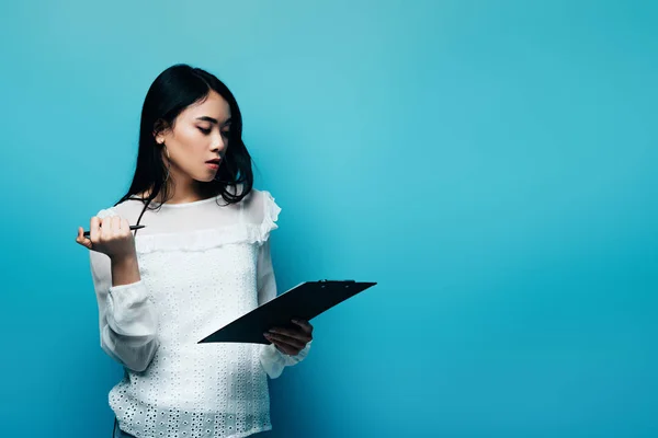 Pensive asian journalist in white blouse holding clipboard on blue background — Stock Photo