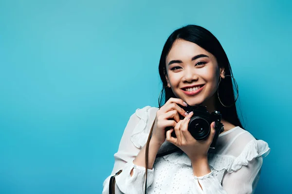 Heureux asiatique femme en blanc chemisier tenant appareil photo numérique sur fond bleu — Photo de stock