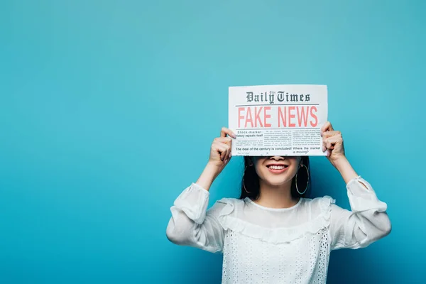 Sonriente asiático mujer en blanco blusa celebración periódico con falso noticias en azul fondo - foto de stock