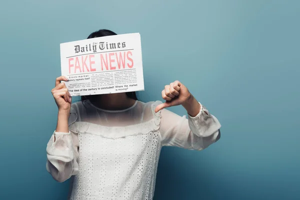 Mujer en blusa blanca sosteniendo periódico con noticias falsas y mostrando el pulgar hacia abajo sobre fondo azul - foto de stock