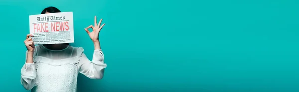 Plan panoramique de femme en chemisier blanc tenant journal avec de fausses nouvelles et montrant ok signe sur fond turquoise — Photo de stock