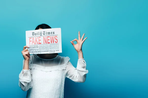 Женщина в белой блузке держит газету с фейковыми новостями и показывает знак ОК на синем фоне — стоковое фото