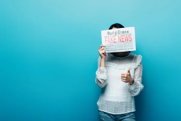 Mujer en blusa blanca sosteniendo periódico con noticias falsas y mostrando el pulgar hacia arriba sobre fondo azul — Stock Photo
