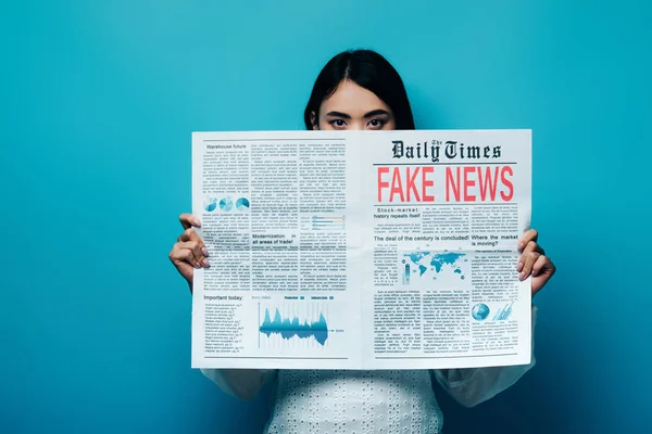 Mujer asiática en blusa blanca sosteniendo periódico con noticias falsas sobre fondo azul — Stock Photo