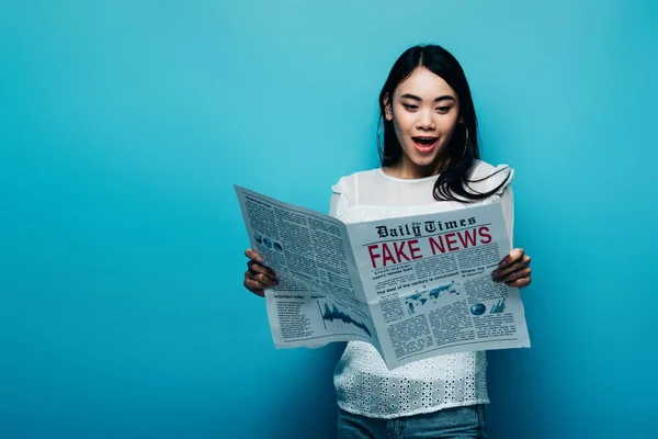 Шокована азіатська жінка в білій блузці читає газету з фальшивими новинами на синьому фоні — стокове фото