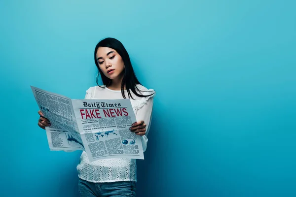 Mujer asiática en blusa blanca leyendo periódico con noticias falsas sobre fondo azul - foto de stock