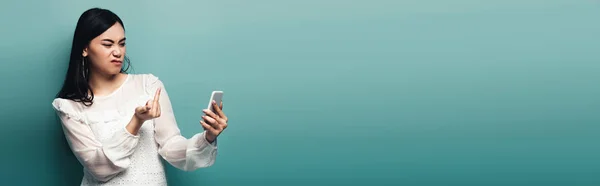Panoramic shot of dissatisfied asian woman in white blouse showing middle finger in smartphone screen on green background — Stock Photo
