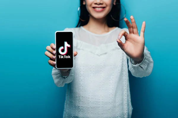 KYIV, UKRAINE - JULY 15, 2019: cropped view of smiling asian woman in white blouse showing ok sign and smartphone with tiktok app on blue background — Stock Photo