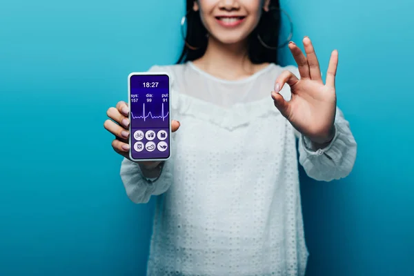 Обрезанный вид улыбающейся азиатской женщины в белой блузке показывает знак ОК и смартфон с приложением здравоохранения на синем фоне — стоковое фото