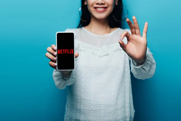 KYIV, UCRÂNIA - JULHO 15, 2019: visão recortada de mulher asiática sorridente em blusa branca mostrando sinal ok e smartphone com o aplicativo netTuber no fundo azul — Fotografia de Stock