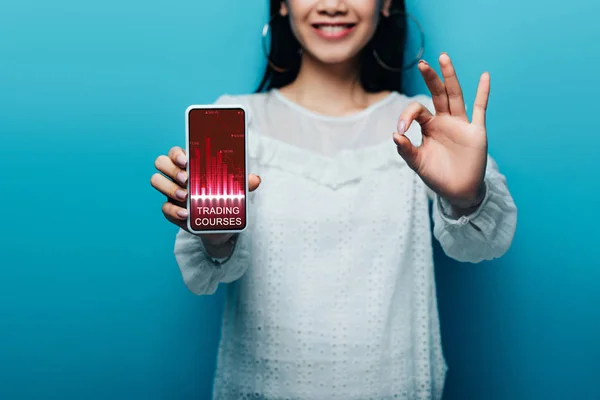 Обрізаний вид усміхненої азіатки в білій блузці, що показує знак ОК і смартфон з додатком на синьому фоні — стокове фото