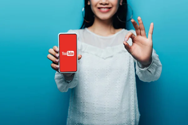 KYIV, UKRAINE - JULY 15, 2019: cropped view of smiling asian woman in white blouse showing ok sign and smartphone with youtube app on blue background — Stock Photo
